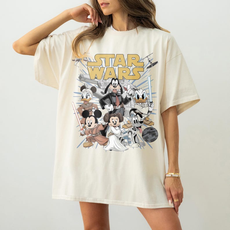May include: Cream-colored t-shirt featuring a graphic of Disney characters in Star Wars attire. The design includes Mickey Mouse, Minnie Mouse, Donald Duck, and Goofy, with the Star Wars logo in gold. The shirt has short sleeves and a relaxed fit.