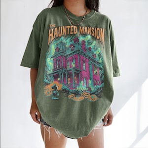 May include: Olive green t-shirt featuring a colorful graphic of a haunted house with the text "THE HAUNTED MANSION". The design includes ghosts, pumpkins, and a cartoon character, perfect for Halloween.