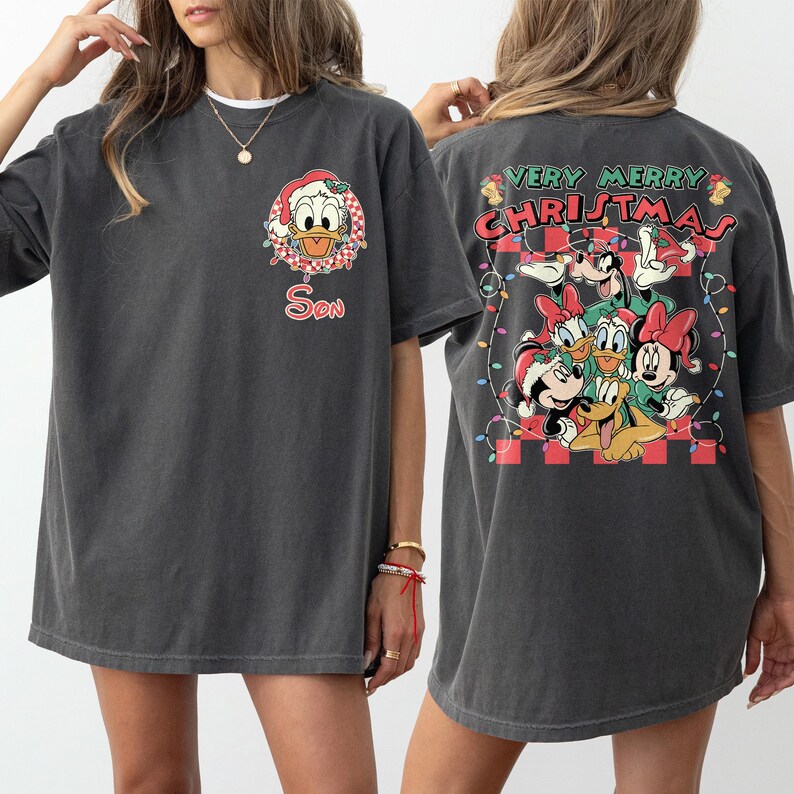 May include: Dark gray t-shirt featuring a Christmas-themed design. The front displays Donald Duck in a Santa hat with the word "Son". The back showcases Mickey, Minnie, Donald, Goofy, and Pluto with the text "Very Merry Christmas".