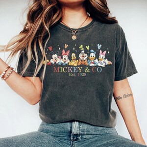 Puede incluir: Camiseta gris oscuro con un gr&aacute;fico colorido de Mickey Mouse y sus amigos. El dise&ntilde;o incluye a Mickey, Minnie, Goofy, Pluto, Donald Duck y Daisy Duck, con el texto "Mickey & Co. Est. 1928".