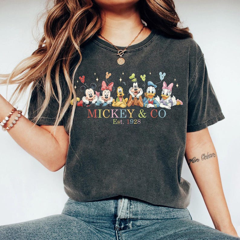 Puede incluir: Camiseta gris oscuro con un gr&aacute;fico colorido de Mickey Mouse y sus amigos. El dise&ntilde;o incluye a Mickey, Minnie, Goofy, Pluto, Donald Duck y Daisy Duck, con el texto "Mickey & Co. Est. 1928".