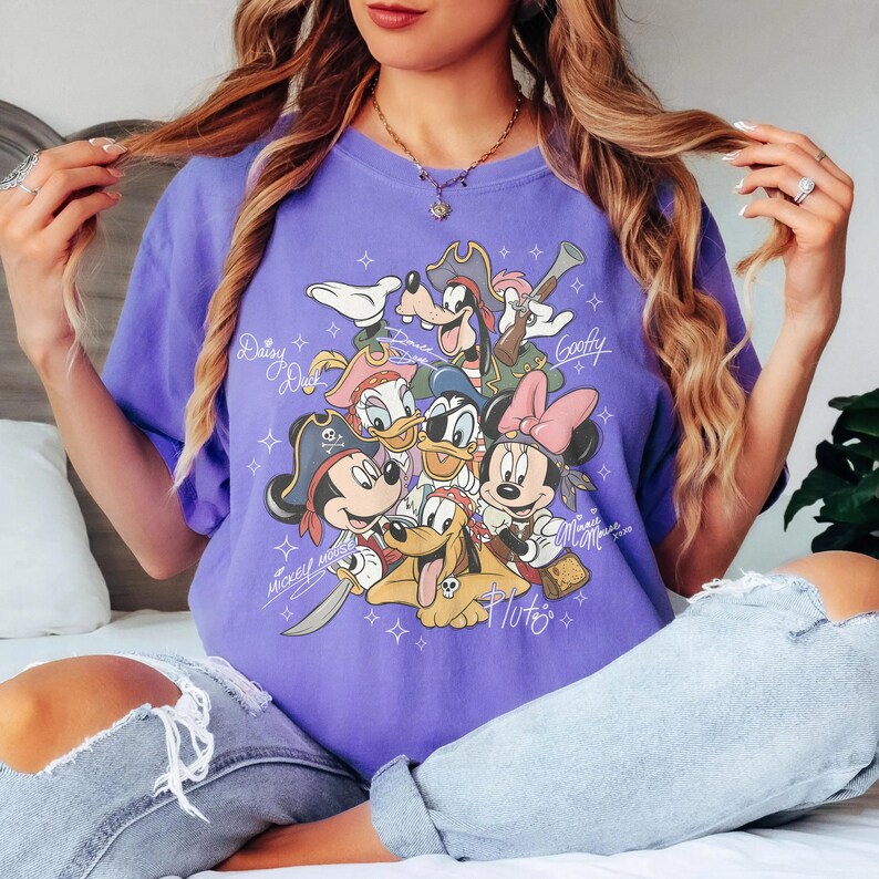 May include: A purple t-shirt featuring a graphic of Disney characters dressed as pirates. The characters include Mickey Mouse, Minnie Mouse, Donald Duck, Daisy Duck, Goofy, and Pluto. The names of the characters are written above or below them in a cursive font.