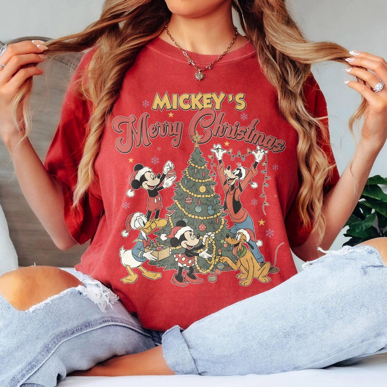 May include: Red t-shirt featuring a festive graphic with Mickey Mouse, Minnie Mouse, Donald Duck, Goofy, and Pluto decorating a Christmas tree. The text "Mickey's Merry Christmas" is displayed above the characters.