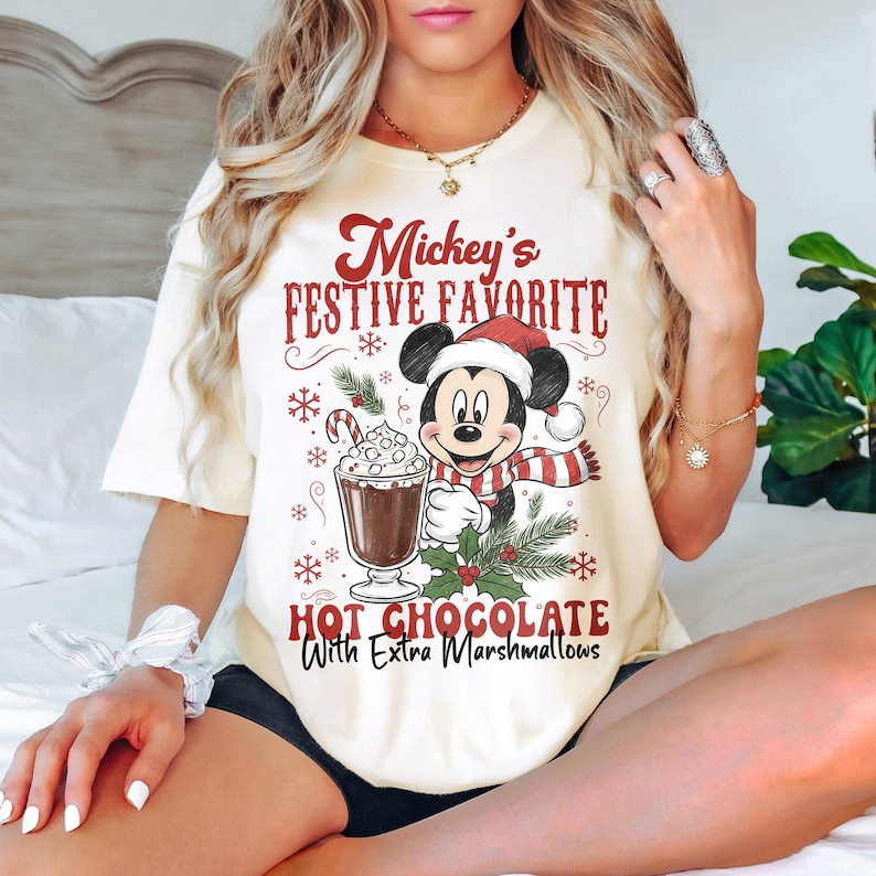 May include: Cream-colored t-shirt featuring Mickey Mouse in a Santa hat and scarf, holding a mug of hot chocolate with marshmallows. The text reads "Mickey's Festive Favorite Hot Chocolate With Extra Marshmallows."
