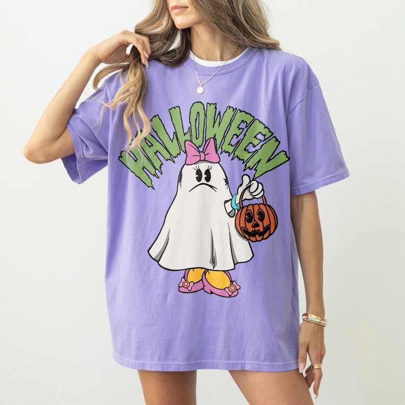 May include: A lavender t-shirt with a Halloween graphic. The graphic features a cartoon ghost character with a pink bow, holding a jack-o'-lantern. The word "HALLOWEEN" is printed above the ghost in green, jagged letters.