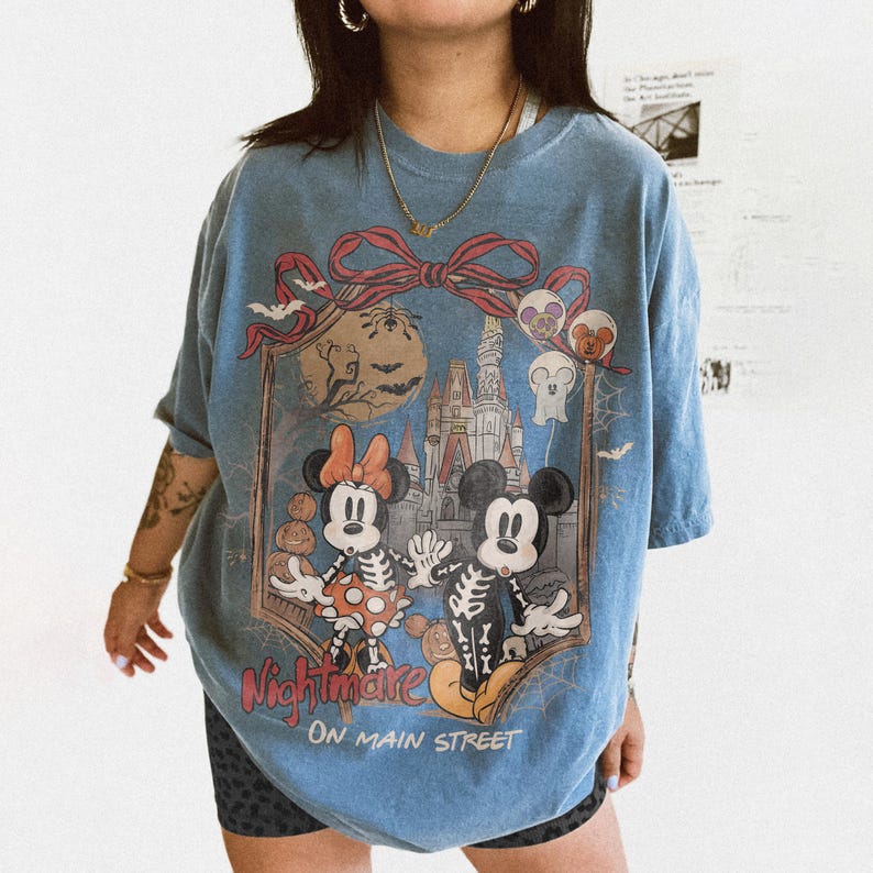 May include: Blue t-shirt featuring a Halloween-themed graphic with Mickey and Minnie Mouse dressed as skeletons in front of a castle. The shirt has the words "Nightmare on Main Street" printed on it.