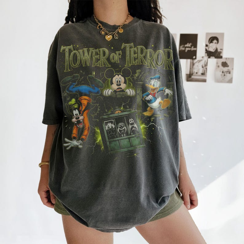 May include: A charcoal gray oversized t-shirt featuring a graphic print of the Tower of Terror with Mickey Mouse, Goofy, and Donald Duck. The design includes green lightning accents and the text Tower of Terror in a stylized font across the chest.