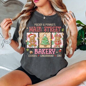 Op de afbeelding: Donkergrijs t-shirt met een feestelijk ontwerp. Het shirt toont de tekst "Mickey & Minnie's Main Street Bakery" met peperkoekkarakters, een kerstboom en de woorden "Cookies, Gingerbread, Cake."