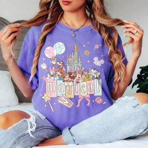 May include: A purple t-shirt featuring a colorful graphic of a castle, Disney characters, and the word "MAGICAL". The design includes Mickey and Minnie Mouse, Donald Duck, and Goofy. The shirt has a relaxed fit.