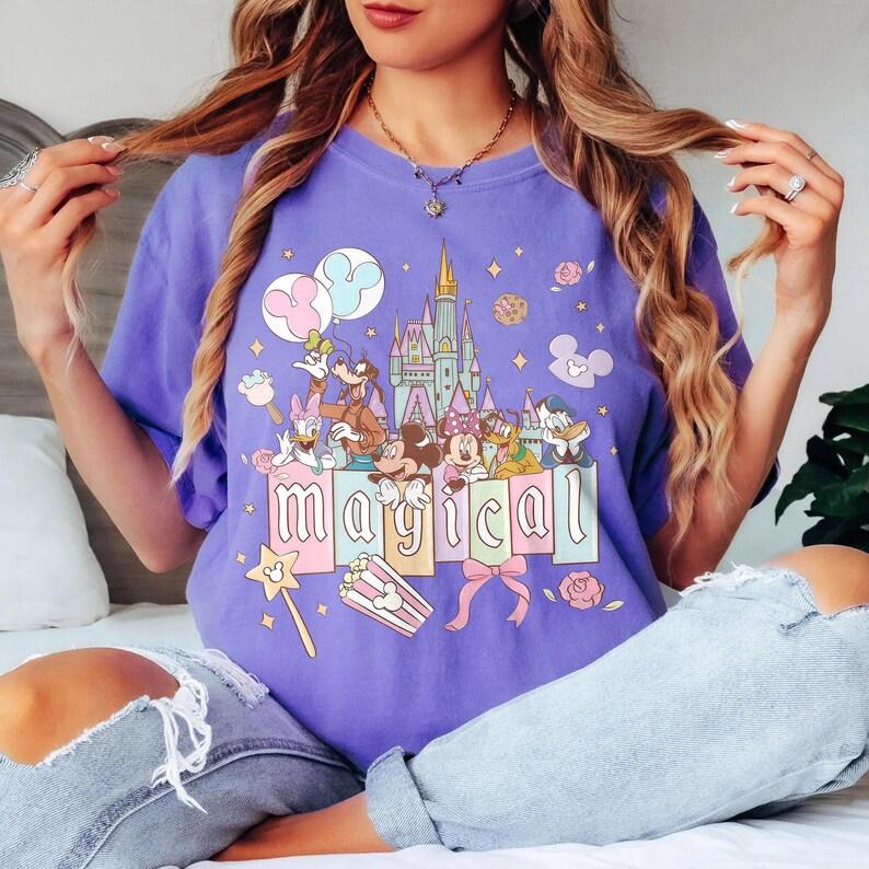 May include: A purple t-shirt featuring a colorful graphic of a castle, Disney characters, and the word "MAGICAL". The design includes Mickey and Minnie Mouse, Donald Duck, and Goofy. The shirt has a relaxed fit.