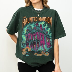 May include: Dark green t-shirt featuring a graphic of a haunted mansion with the text "THE HAUNTED MANSION". The graphic includes a pink house, pumpkins, and ghostly green flames. The shirt is a relaxed fit.
