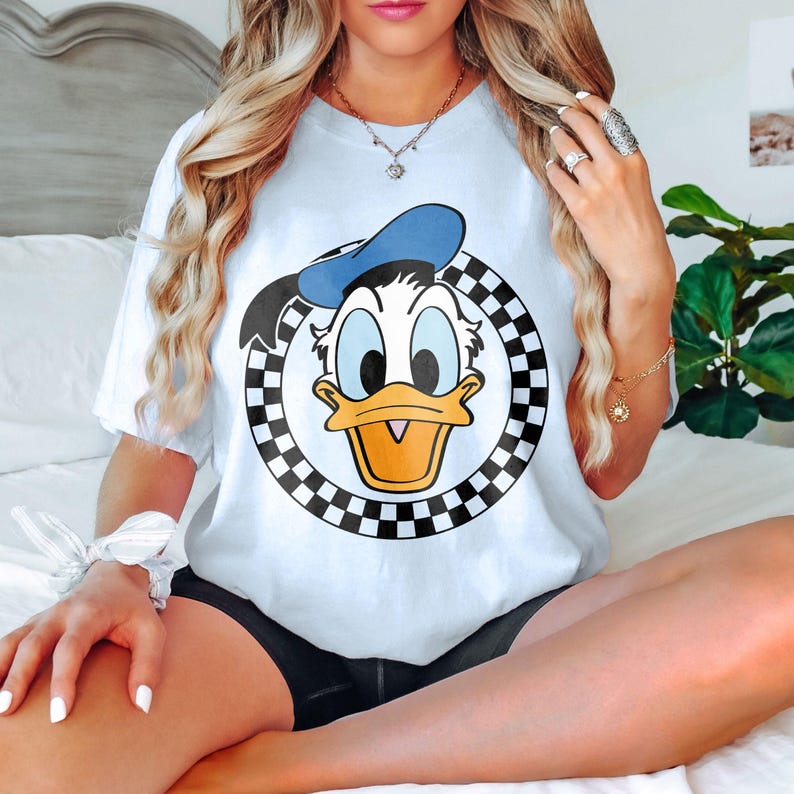 May include: White t-shirt with a graphic of Donald Duck in a black and white chequered circle. Donald Duck is wearing a blue hat and has a yellow beak. The shirt is short-sleeved.