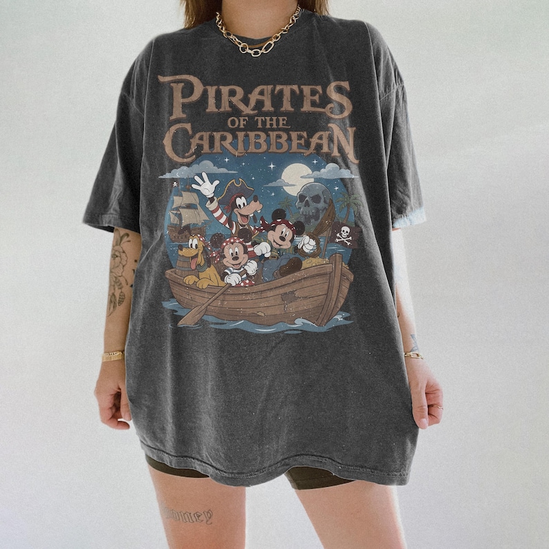 K&ouml;nnte beinhalten: Dunkelgraues T-Shirt mit einem Aufdruck von Disney-Figuren in einem Boot und dem Text "Pirates of the Caribbean". Das Design umfasst Micky Maus, Goofy und Donald Duck. Das Shirt hat einen Vintage-Look.