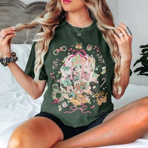 May include: Dark green t-shirt featuring a festive Disney design. The graphic includes Mickey and Minnie Mouse, Donald and Daisy Duck, and Goofy, with a castle backdrop and holiday accents like bows, gifts, and candy canes. The shirt is a relaxed fit.