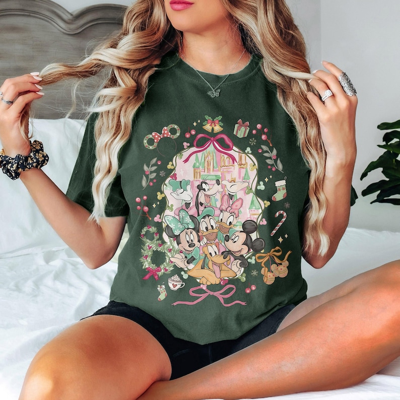 May include: Dark green t-shirt featuring a festive Disney design. The graphic includes Mickey and Minnie Mouse, Donald and Daisy Duck, and Goofy, with a castle backdrop and holiday accents like bows, gifts, and candy canes. The shirt is a relaxed fit.