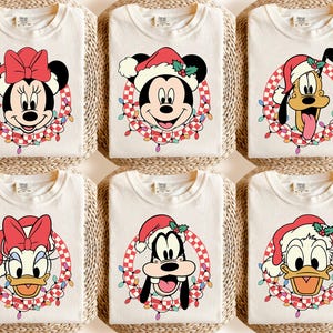 May include: Six cream-colored t-shirts featuring Disney characters in Santa hats and festive wreaths. Minnie Mouse, Mickey Mouse, Pluto, Donald Duck, Goofy, and Daisy Duck are depicted. Each shirt has a unique character design, perfect for holiday celebrations.