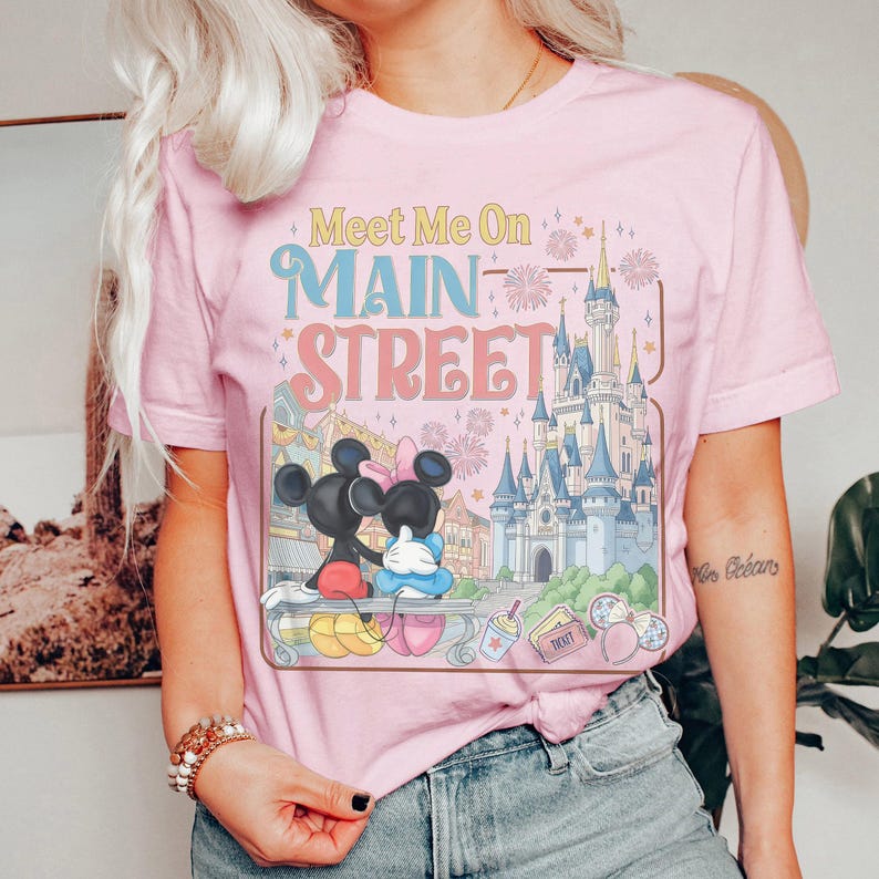 Op de afbeelding: Roze T-shirt met de tekst "Meet Me On Main Street" in pastelkleuren. De afbeelding toont Mickey en Minnie Mouse, een kasteel en vuurwerk. Het shirt heeft een ronde hals en korte mouwen.