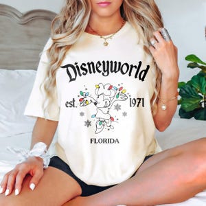 May include: Cream-coloured t-shirt featuring the text "Disneyworld" in a black, stylised font. Below is a graphic of Mickey Mouse with Christmas lights, the text "est. 1971", and the word "FLORIDA".