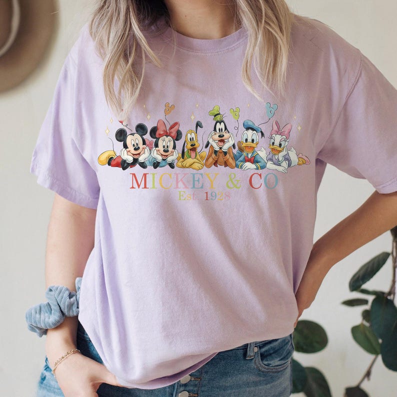 Puede incluir: Camiseta lila claro con un gr&aacute;fico colorido de Mickey Mouse, Minnie Mouse, Pluto, Goofy, Donald Duck y Daisy Duck. El texto "Mickey & Co. Est. 1928" est&aacute; impreso debajo. La camiseta tiene mangas cortas.