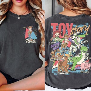 K&ouml;nnte beinhalten: Dunkelgraues T-Shirt mit einer farbenfrohen Grafik von Toy Story-Charakteren, darunter Woody, Buzz Lightyear und Rex. Auf der Vorderseite des Shirts befindet sich eine kleinere Grafik von Woody mit dem Wort "Uncle". Auf der R&uuml;ckseite des Shirts stehen die Worte "Toy Story" in einem farbenfrohen Design.