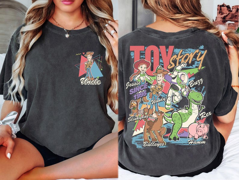 K&ouml;nnte beinhalten: Dunkelgraues T-Shirt mit einer farbenfrohen Grafik von Toy Story-Charakteren, darunter Woody, Buzz Lightyear und Rex. Auf der Vorderseite des Shirts befindet sich eine kleinere Grafik von Woody mit dem Wort "Uncle". Auf der R&uuml;ckseite des Shirts stehen die Worte "Toy Story" in einem farbenfrohen Design.