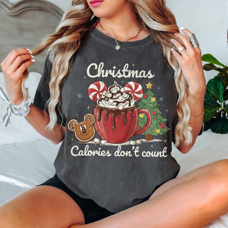 May include: Dark gray t-shirt with a Christmas-themed graphic. The design features a red mug of hot chocolate with Mickey Mouse ears, candy canes, marshmallows, and a gingerbread cookie. Text reads "Christmas Calories don't count."