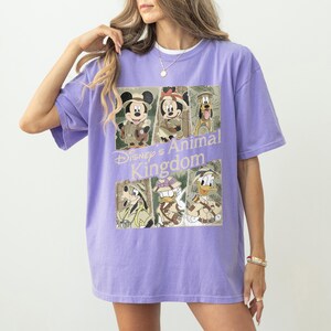May include: A lilac-colored t-shirt featuring a graphic of Disney characters in safari attire. The design includes Mickey Mouse, Minnie Mouse, Pluto, Goofy, and Donald Duck, with the text "Disney's Animal Kingdom" in a stylized font.