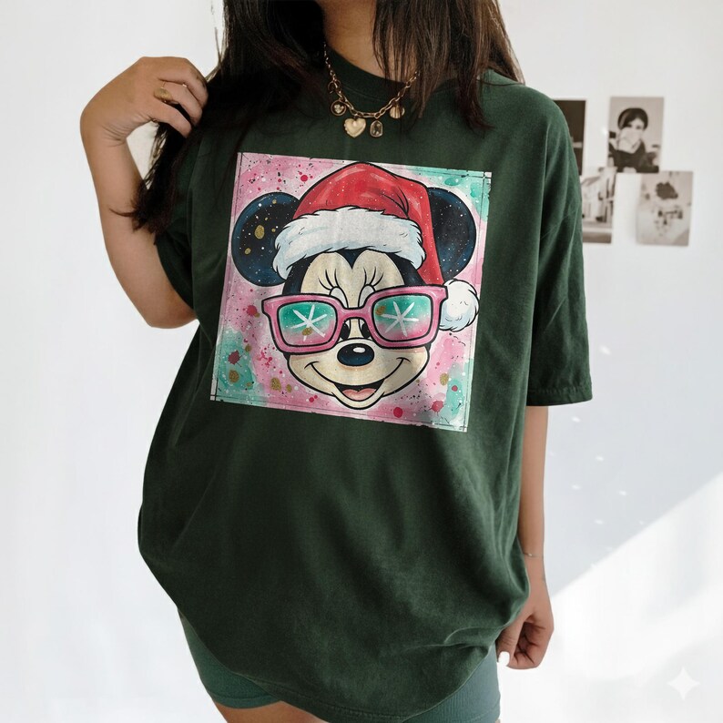 May include: Dark green t-shirt featuring a graphic of Mickey Mouse wearing a Santa hat and pink sunglasses. The graphic is set against a colorful, watercolor-style background. The shirt has short sleeves and a relaxed fit.