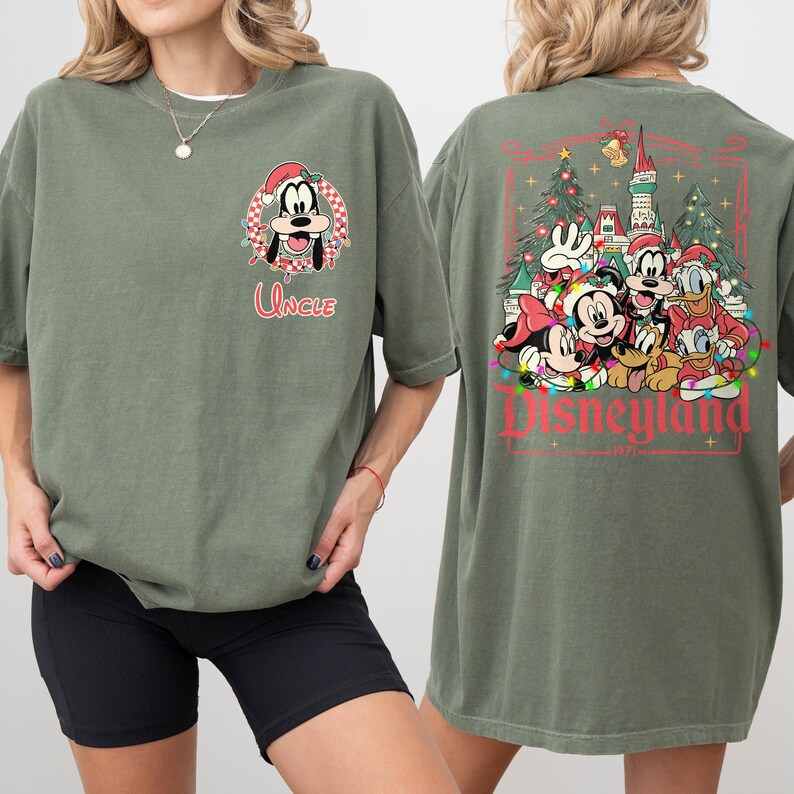 K&ouml;nnte beinhalten: Olivgr&uuml;nes T-Shirt mit festlichem Design. Auf der Vorderseite ist ein Cartoon von Goofy mit Weihnachtsm&uuml;tze und dem Wort "Uncle" zu sehen. Auf der R&uuml;ckseite sind Micky, Minnie, Donald und Goofy vor einem Schloss mit dem Wort "Disneyland" und dem Jahr "1971" abgebildet.
