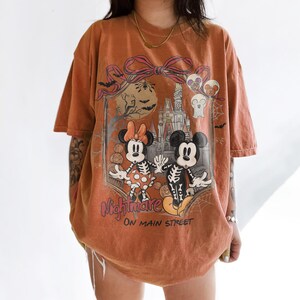 May include: Orange t-shirt featuring a Halloween-themed graphic with Mickey and Minnie Mouse in skeleton costumes. The design includes a castle, bats, and the text "Nightmare on Main Street."