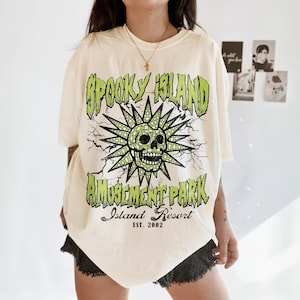 K&ouml;nnte beinhalten: Cremefarbenes T-Shirt mit einem gr&uuml;nen Aufdruck mit der Aufschrift "Spooky Island Amusement Park Island Resort Est. 2002". Die Grafik zeigt einen Totenkopf mit Sonnenstrahlen und Blitzen.