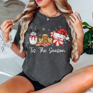 May include: Dark gray t-shirt with a festive graphic. The design includes a jar of milk with Mickey Mouse ears, cookies, and a candy cane Santa. The text "Tis The Season" is written below the graphic.