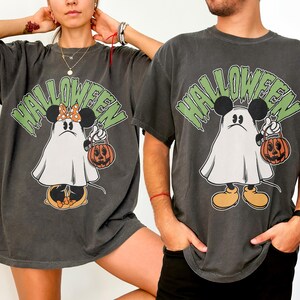 May include: Dark gray Halloween t-shirts featuring a cartoon ghost character with Mickey Mouse ears holding a jack-o'-lantern. The word "HALLOWEEN" is printed above the character in green. The shirts have a vintage, distressed look.