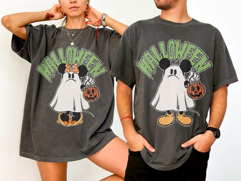 May include: Dark gray Halloween t-shirts featuring a cartoon ghost character with Mickey Mouse ears holding a jack-o'-lantern. The word "HALLOWEEN" is printed above the character in green. The shirts have a vintage, distressed look.