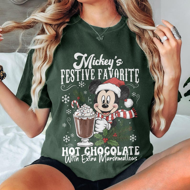 May include: Dark green t-shirt featuring Mickey Mouse in a Santa hat and scarf, holding a hot chocolate with marshmallows. The text reads "Mickey's Festive Favorite Hot Chocolate With Extra Marshmallows."