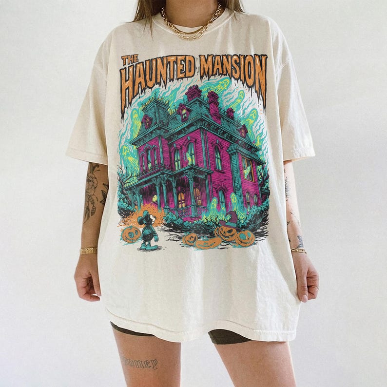 May include: Cream-colored t-shirt featuring a graphic of a haunted house with the text "The Haunted Mansion." The design includes a pink and teal house, ghosts, pumpkins, and a cartoon character.
