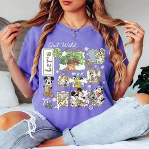 May include: A purple t-shirt with the text "Get Wild" and "Let's" printed on it. The shirt features cartoon characters in safari outfits, including Mickey Mouse, Donald Duck, and Chip and Dale. The design includes a tree and other safari-themed elements.