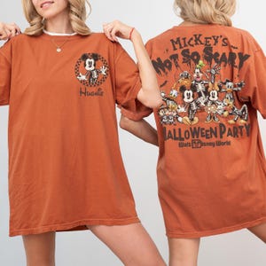 May include: Burnt orange t-shirt featuring a Halloween-themed Mickey Mouse skeleton graphic on the front and back. The front has the name "Hughie" and the back says "Mickey's Not So Scary Halloween Party" with Disney characters.