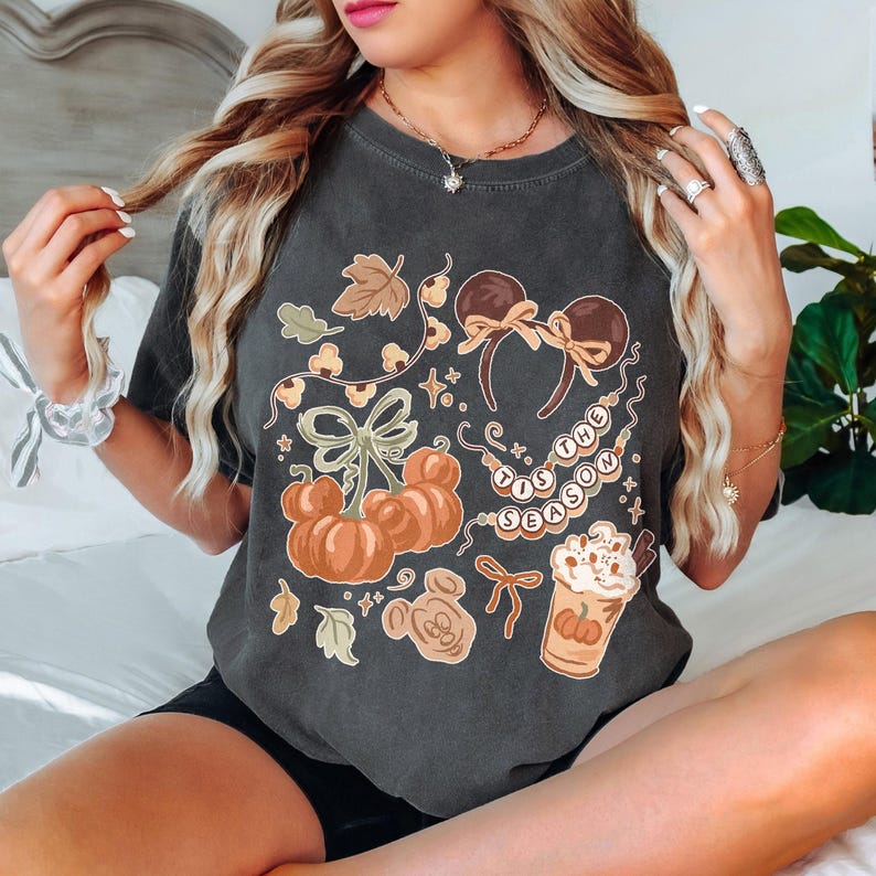 May include: A charcoal gray t-shirt featuring a graphic print of autumn-themed icons including pumpkins, mouse-ear headbands, a whipped cream drink, and a string of beads with the text 'TIS THE SEASON' alongside various leaves and bows.