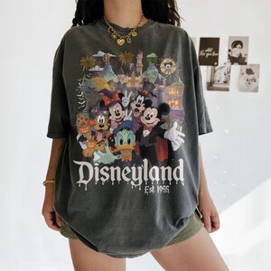 May include: A charcoal gray short-sleeve t-shirt featuring a colorful Halloween-themed graphic of Mickey Mouse, Minnie Mouse, Donald Duck, and Pluto in front of a castle. The text Disneyland Est. 1955 is printed in white below the characters.