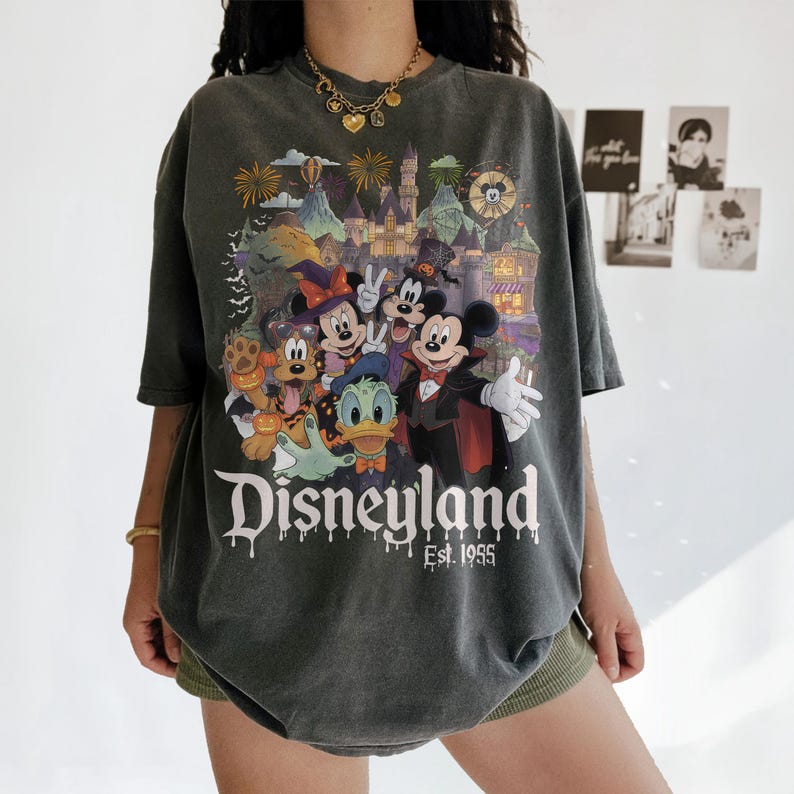 May include: A charcoal gray short-sleeve t-shirt featuring a colorful Halloween-themed graphic of Mickey Mouse, Minnie Mouse, Donald Duck, and Pluto in front of a castle. The text Disneyland Est. 1955 is printed in white below the characters.