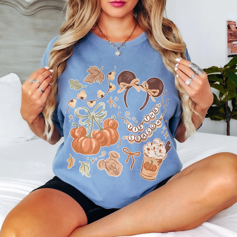 May include: A blue crewneck t-shirt featuring a graphic print of orange pumpkins, mouse-ear headbands with bows, a string of beads with the text 'TIS THE SEASON', and a whipped cream topped beverage, all accented with small leaves and stars.