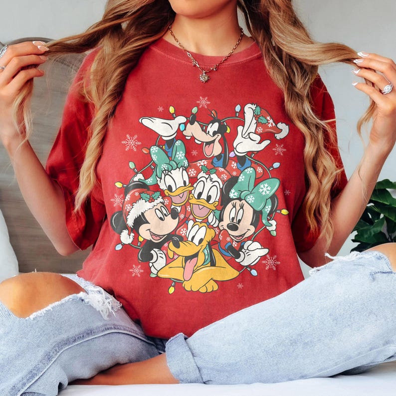 May include: Red t-shirt featuring a festive design with Disney characters, including Mickey and Minnie Mouse, Donald and Daisy Duck, Goofy, and Pluto, surrounded by Christmas lights and snowflakes. The shirt has short sleeves and a crew neck.