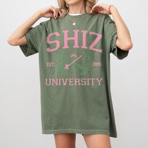 Pu&ograve; includere: T-shirt verde oliva con la scritta rosa "SHIZ UNIVERSITY". La maglietta presenta un design a stemma con la scritta "EST. 1995". La maglietta &egrave; in stile casual e oversize.
