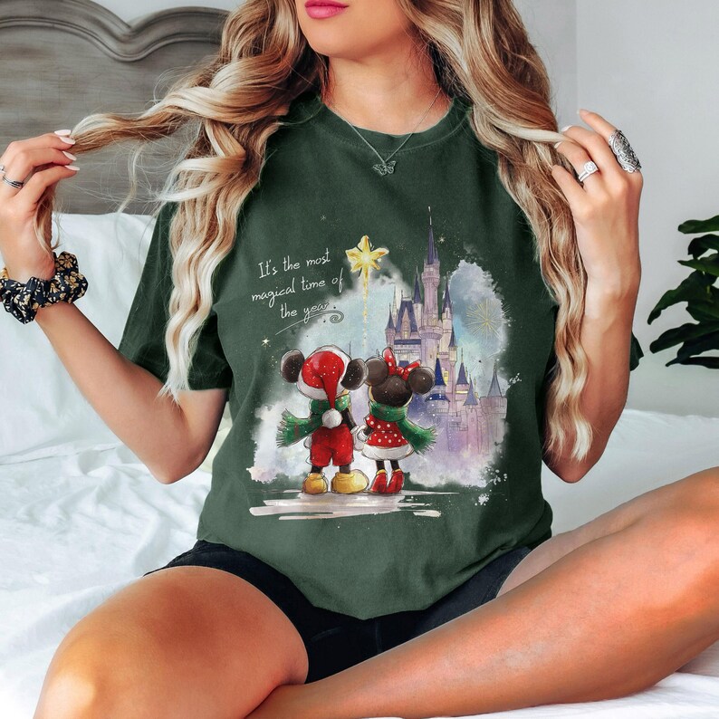 Pu&ograve; includere: T-shirt verde scuro con un'illustrazione festiva di Topolino e Minnie Mouse in abiti natalizi, davanti a un castello. La maglietta include il testo "It's the most magical time of the year."