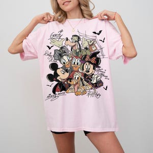 Może przedstawiać: Jasnor&oacute;żowy t-shirt z nadrukiem graficznym przedstawiającym postacie Disneya w kostiumach na Halloween, w tym Myszkę Miki, Myszkę Minnie, Goofy'ego, Kaczorkę Daisy i Pluto. Wz&oacute;r zawiera pajęczyny, nietoperze oraz imiona postaci napisane kursywą.