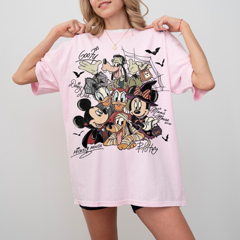 Może przedstawiać: Jasnor&oacute;żowy t-shirt z nadrukiem graficznym przedstawiającym postacie Disneya w kostiumach na Halloween, w tym Myszkę Miki, Myszkę Minnie, Goofy'ego, Kaczorkę Daisy i Pluto. Wz&oacute;r zawiera pajęczyny, nietoperze oraz imiona postaci napisane kursywą.