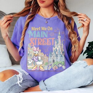 K&ouml;nnte beinhalten: Ein lila T-Shirt mit dem Text "Meet Me On Main Street" und einer Grafik von Daisy Duck vor einem Schloss und Geb&auml;uden. Das Design beinhaltet Feuerwerk und andere Details im Vintage-Stil.