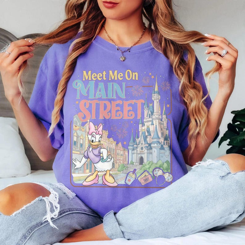 K&ouml;nnte beinhalten: Ein lila T-Shirt mit dem Text "Meet Me On Main Street" und einer Grafik von Daisy Duck vor einem Schloss und Geb&auml;uden. Das Design beinhaltet Feuerwerk und andere Details im Vintage-Stil.