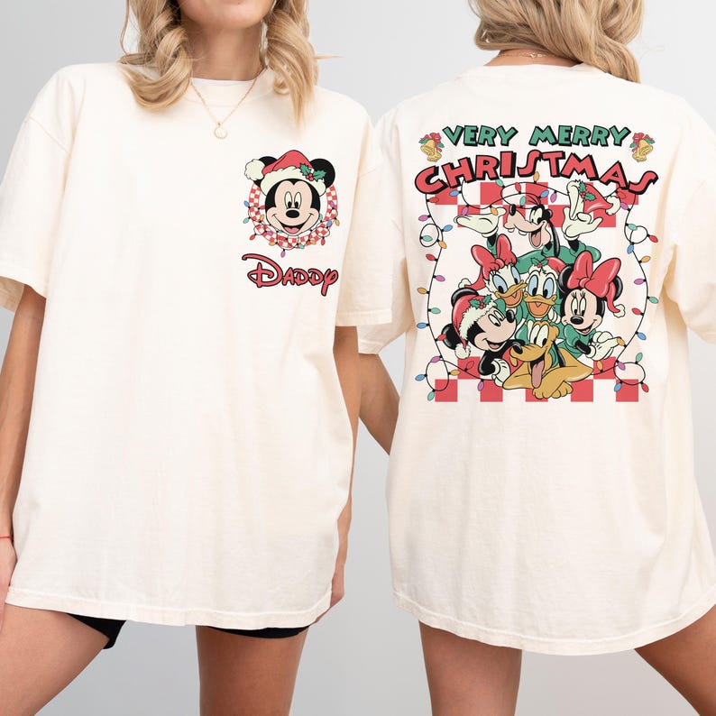 May include: Cream-colored t-shirt featuring a Christmas-themed Mickey Mouse design on the front and a group of Disney characters with the text "VERY MERRY CHRISTMAS" on the back. The design includes festive elements like Santa hats and string lights.